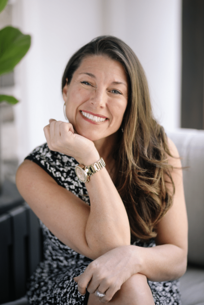 A woman in a stylish dress rests her chin on her hand, showcasing long, flowing hair and a gold watch against a soft, blurred background.