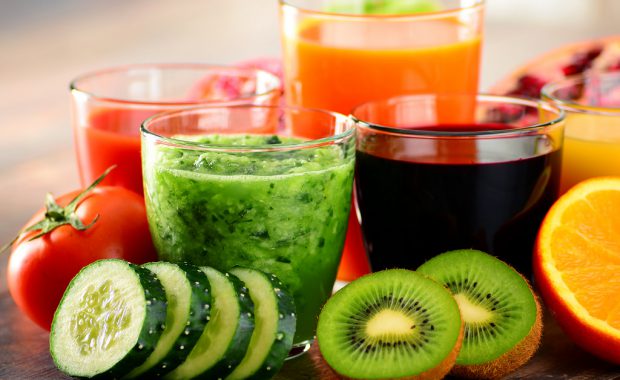 Various glasses of fruit and vegetable juices with sliced cucumber, kiwi, and an orange half in the foreground.
